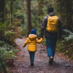 Adult and child walking hand-in-hand through a forest trail, symbolizing modeling, guidance, and learning through observation.